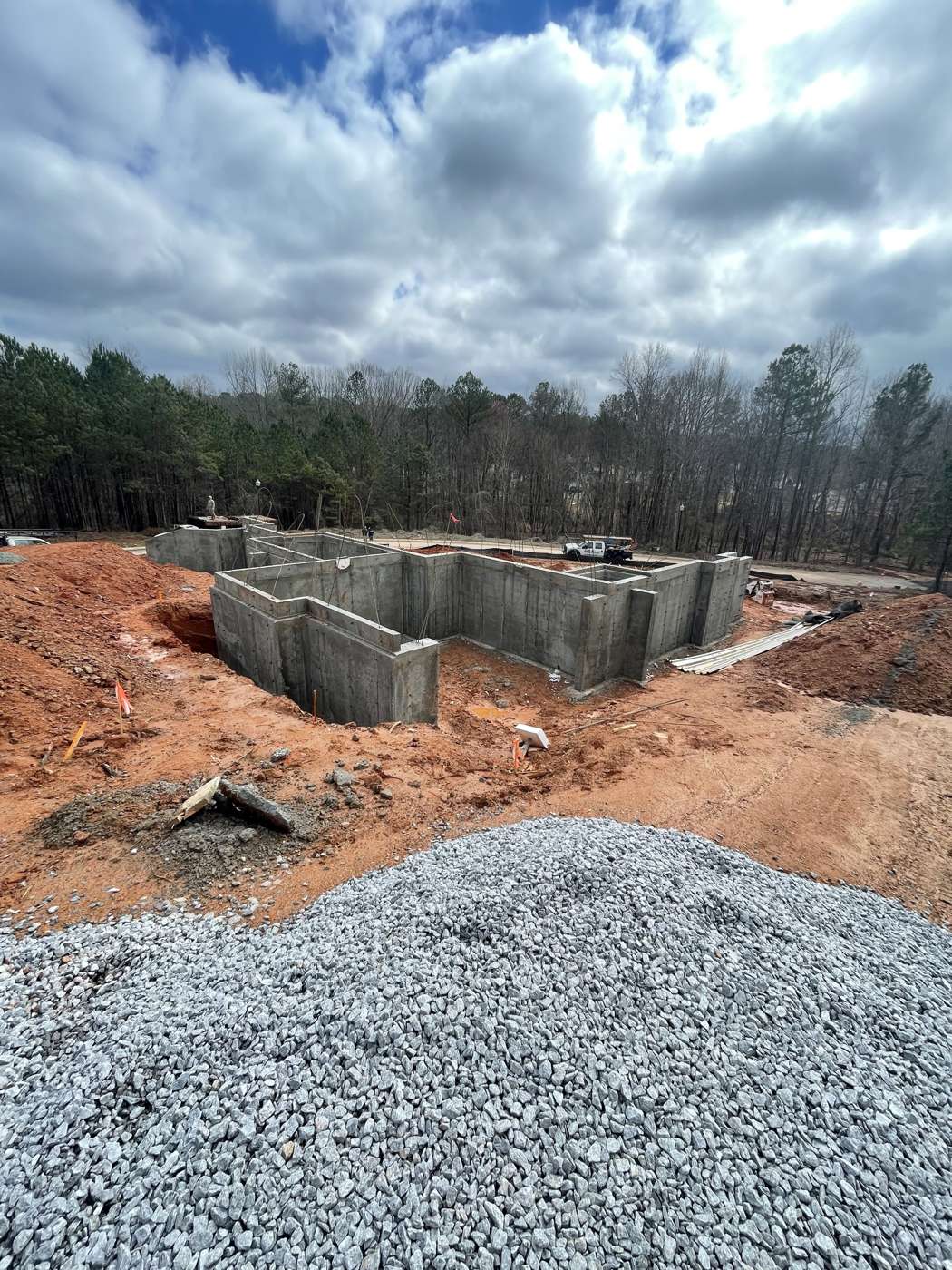 Poured concrete foundation walls on a red clay site behind a pile of gravel