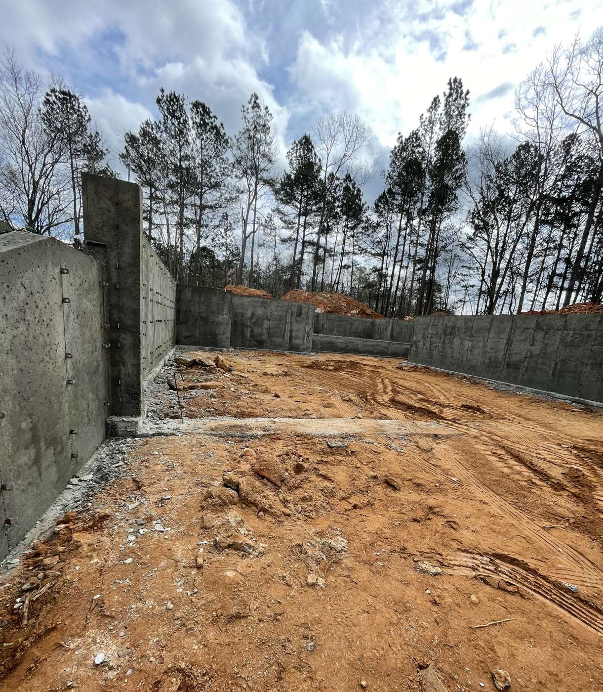 Tall poured concrete wall along an excavated red clay lot with pines above